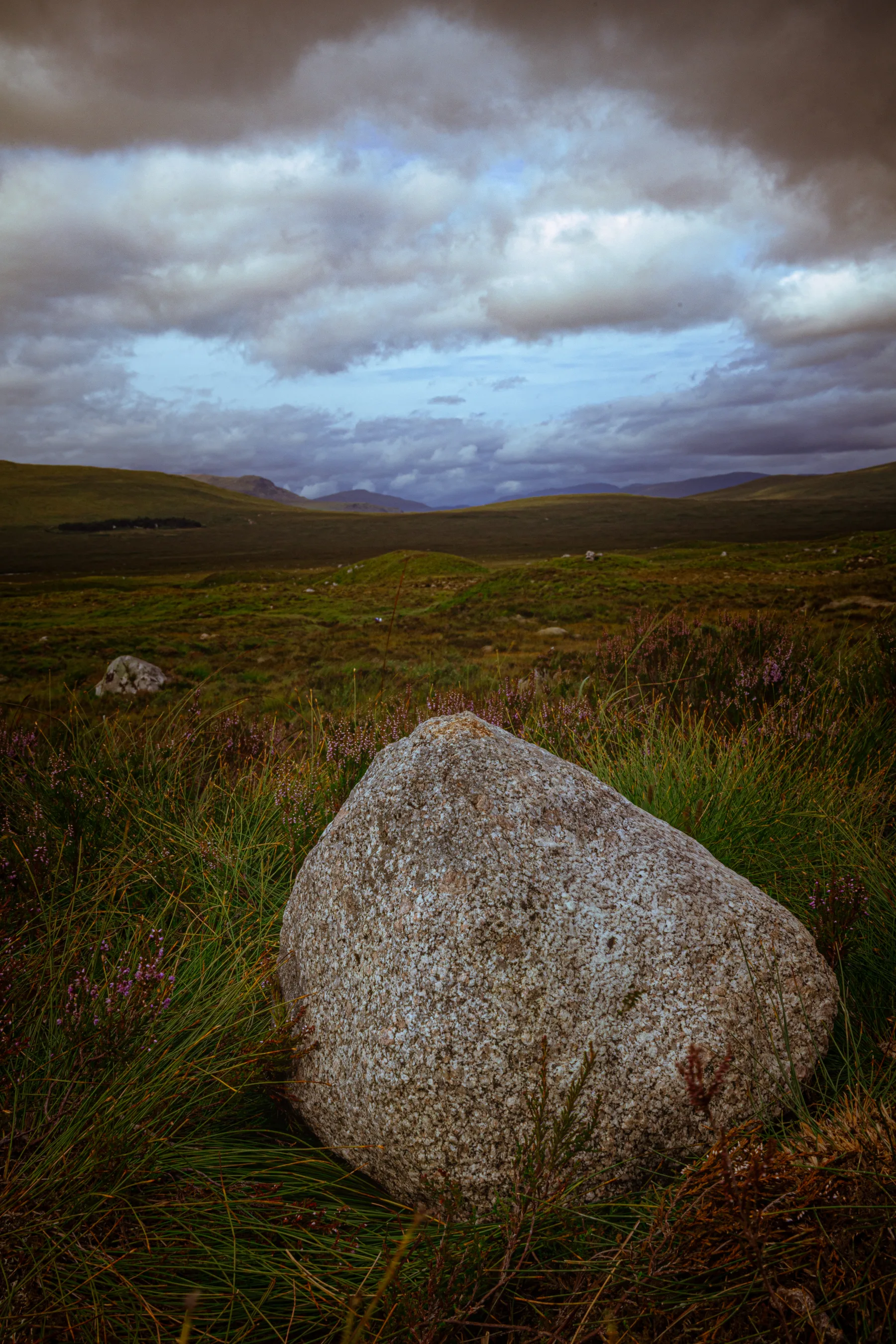 Large granite boulder in grassy moorland with rolling hills and dramatic cloudy sky in the distance.