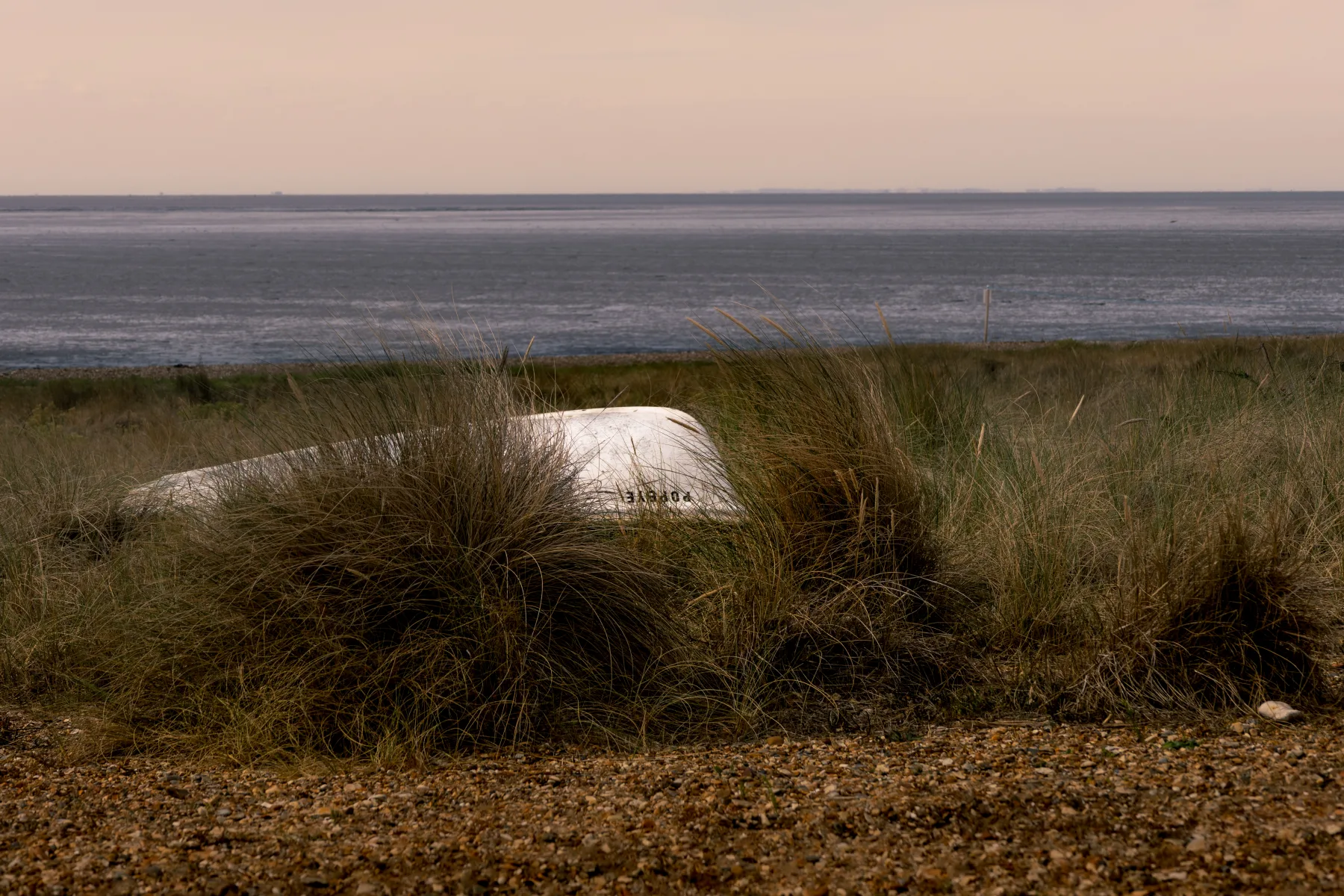 White boat hull resting in dune grass with calm sea and pale pink sky on the horizon.