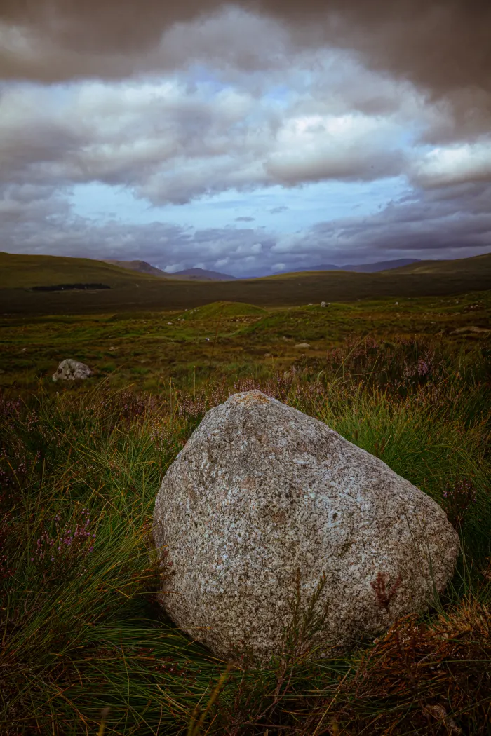 Large granite boulder in grassy moorland with rolling hills and dramatic cloudy sky in the distance.