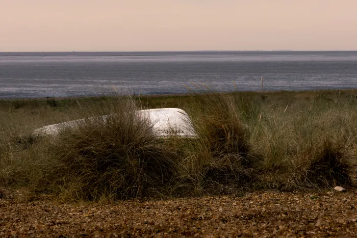 White boat hull resting in dune grass with calm sea and pale pink sky on the horizon.