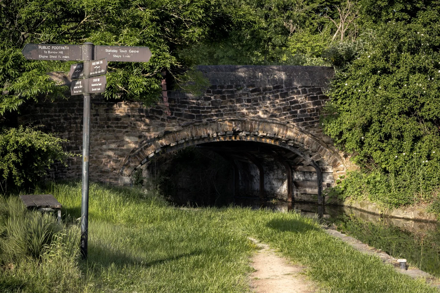 Wayfinding sign beside a grassy towpath leading into a stone bridge tunnel over a narrow canal.