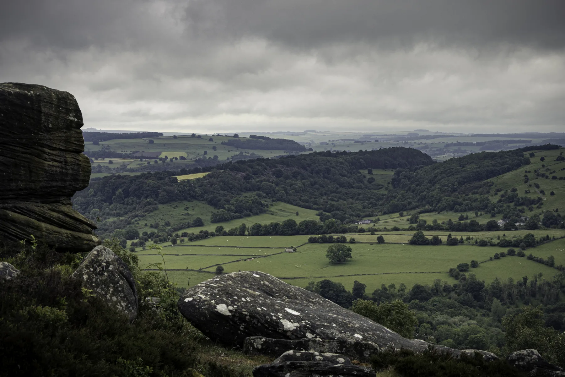 Rocky outcrop overlooking green patchwork fields and wooded hills under a cloudy sky