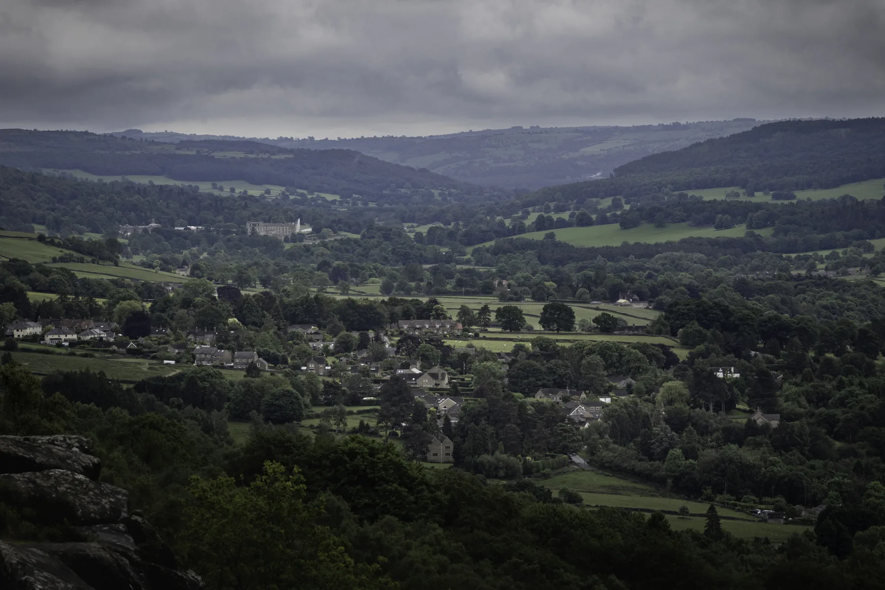 Village nestled in a green valley with rolling hills and patchwork fields under a cloudy sky