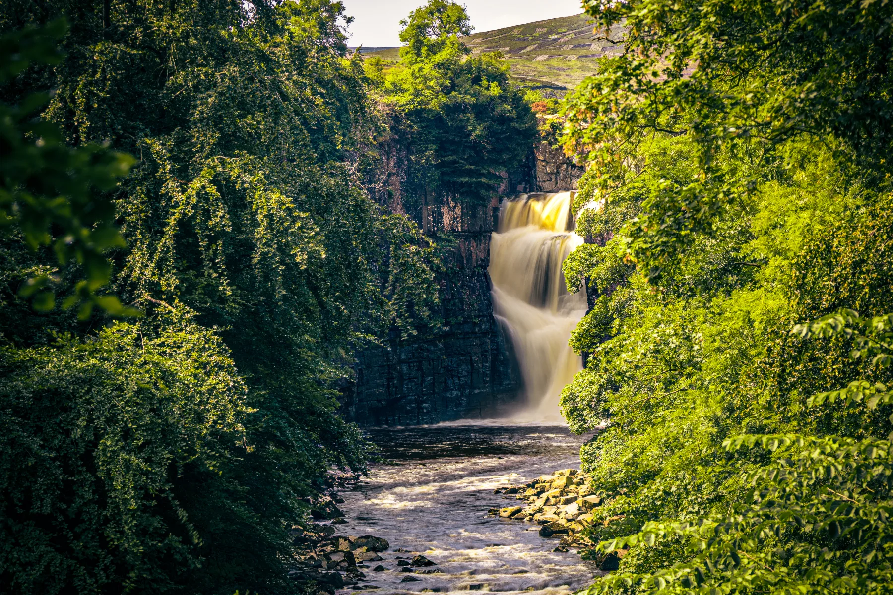 Waterfall cascading into a rocky river, framed by dense green trees with hills in the background.