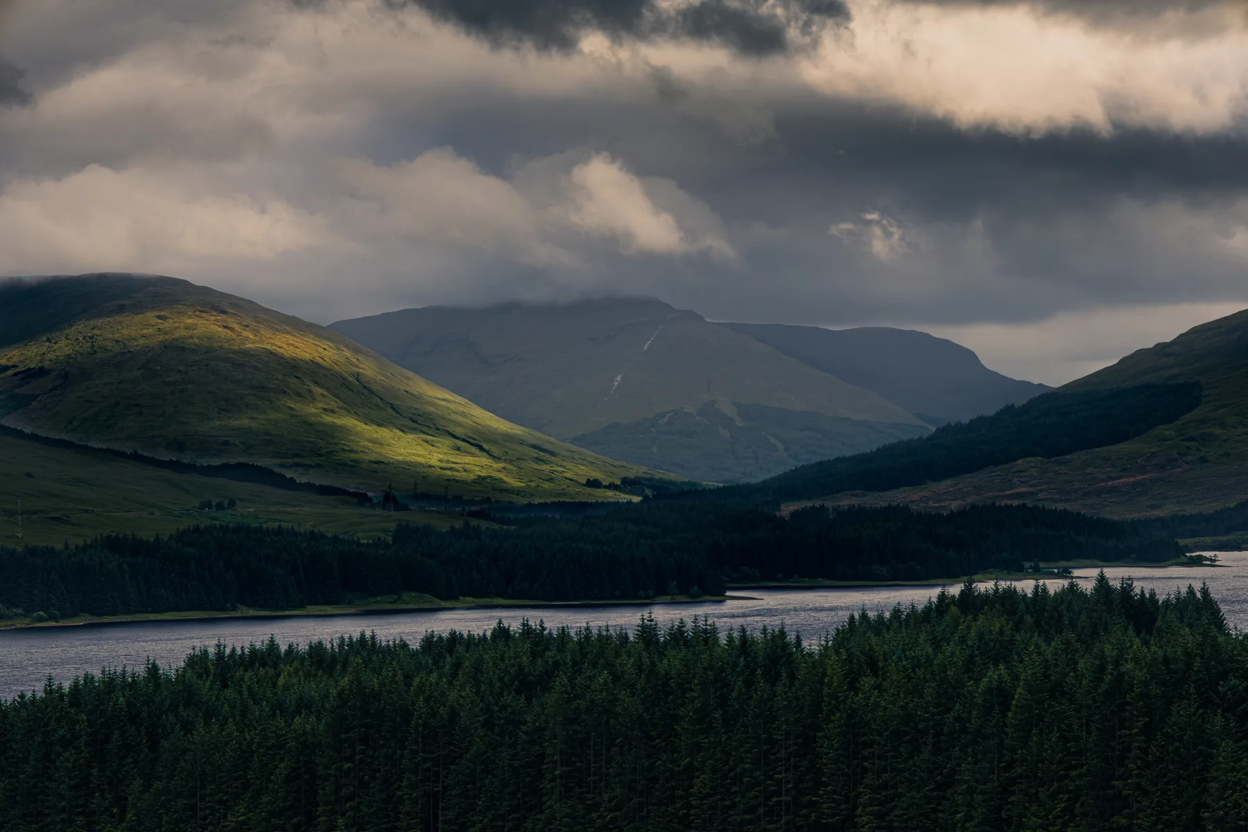 Sunlit green hills and dark forest beside a lake under heavy clouds, with distant mountains in shadow