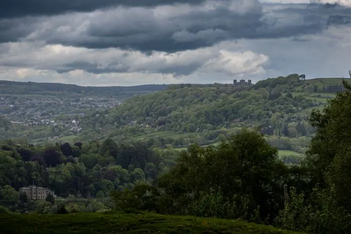 Rolling green hills and valley town under dark storm clouds, with distant hilltop ruins on the horizon.