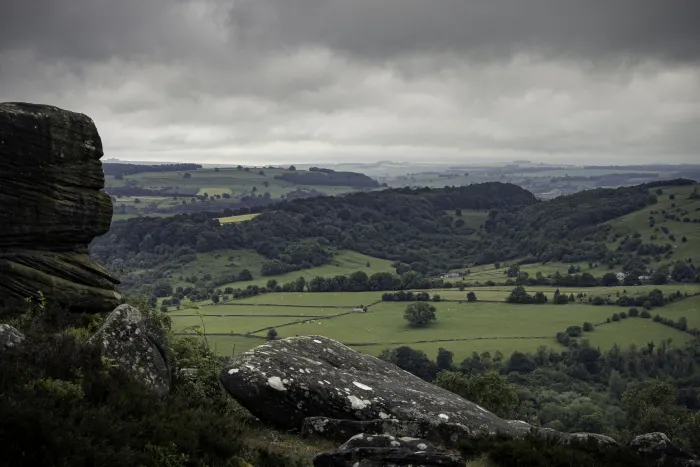 Rocky outcrop overlooking green patchwork fields and wooded hills under a cloudy sky