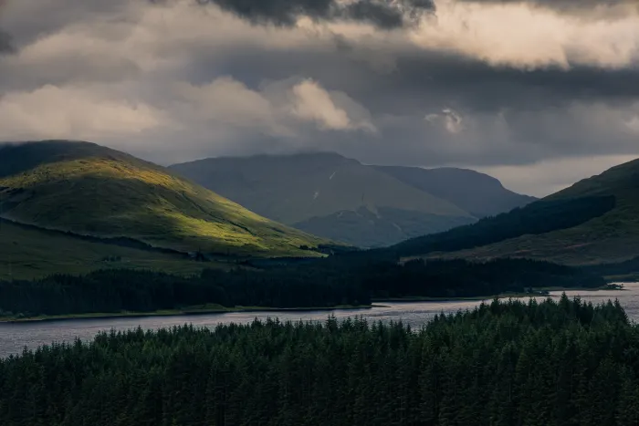 Sunlit green hills and dark forest beside a lake under heavy clouds, with distant mountains in shadow
